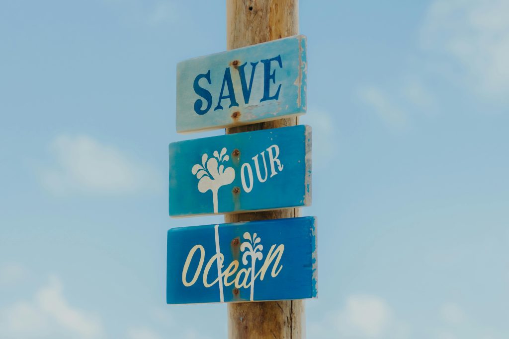 A weathered wooden sign on a beach reads 'Save Our Ocean' against a clear blue sky.